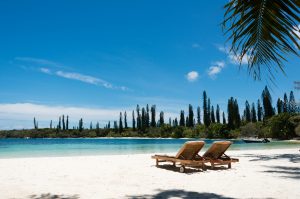 Kanumera beach on the Isle of Pines in New Caledonia. There are a pair of beachchairs on the white sand. The bay is surrounded by pacific cedars and is known as one of the most beautiful bays in the world.